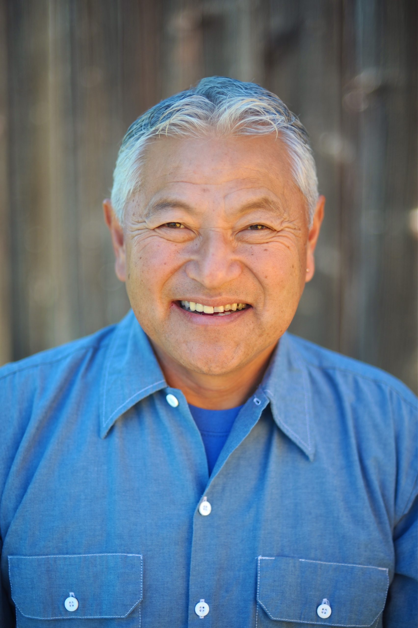 A man wearing a blue shirt smiles standing in front of a brown background